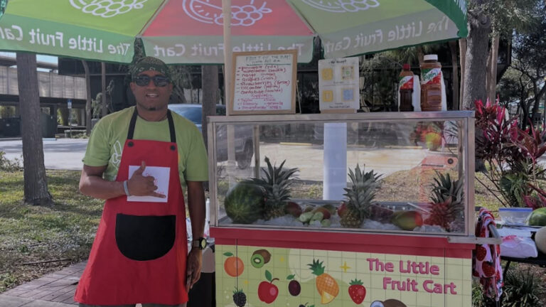 a person stands next to a fruit cart. He's wearing a red apron. A colorful umbrella provides shade